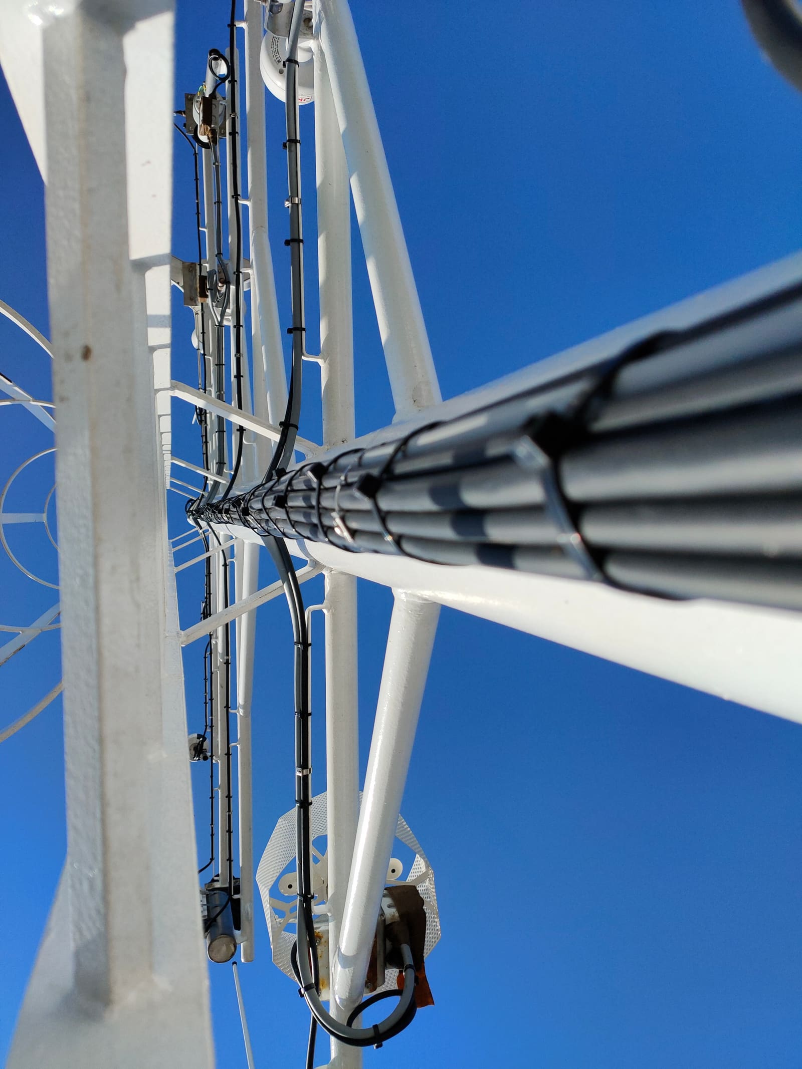 Externally routed cable bundle climbing a navigation tower's lattice mast under a clear blue sky