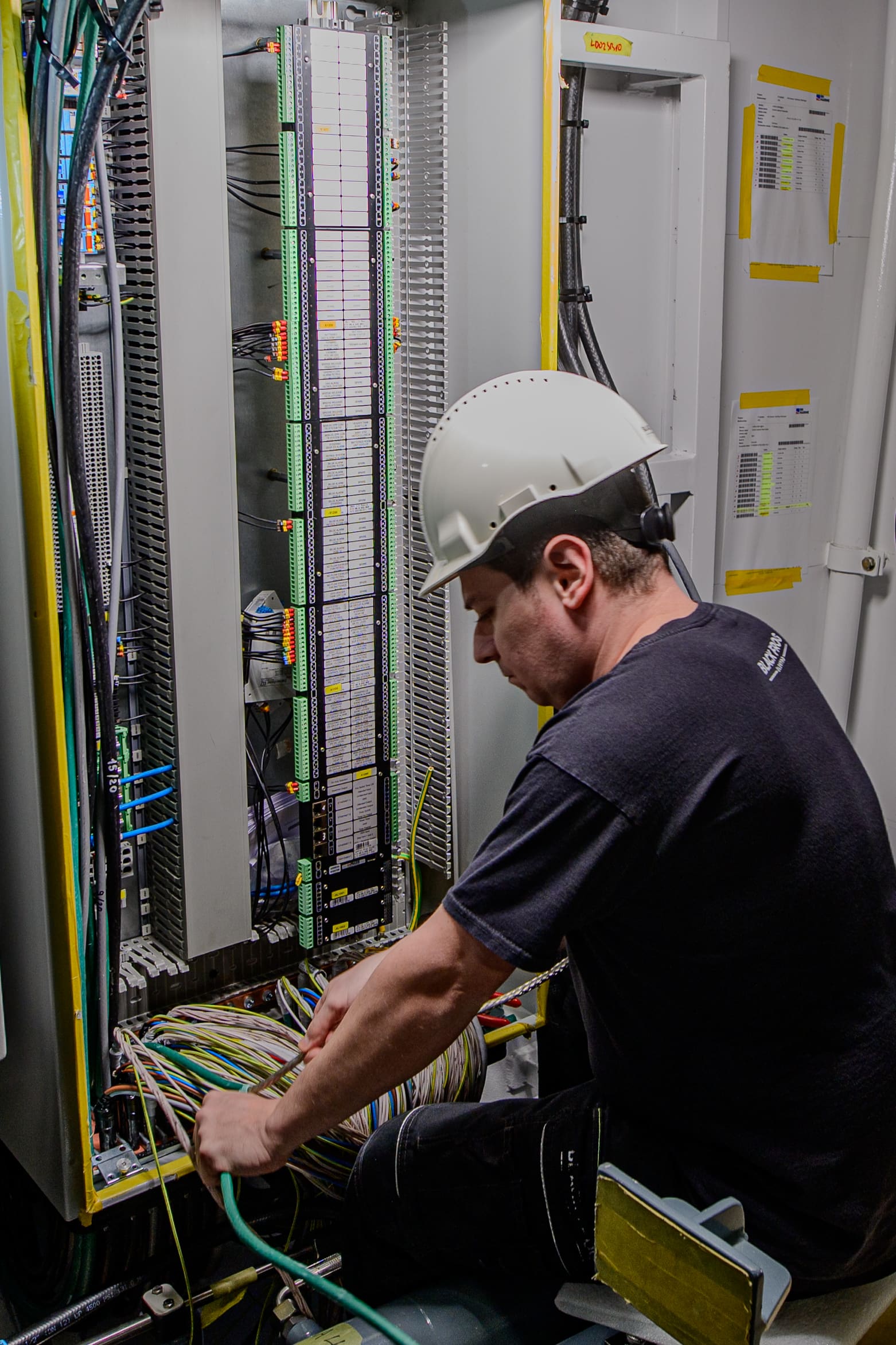 Marine electrical technician in white hardhat terminating control cables into a tall PLC marshalling cabinet aboard a vessel