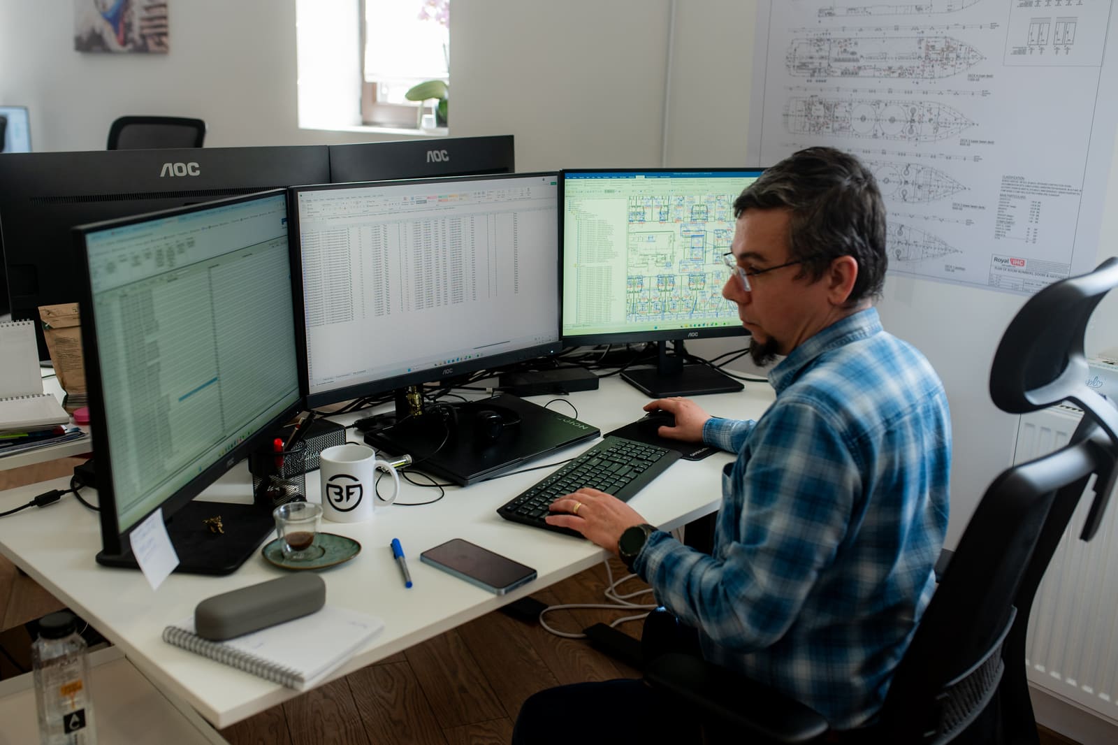 Marine electrical engineer working on vessel technical schematics across three monitors with drawings pinned on the wall