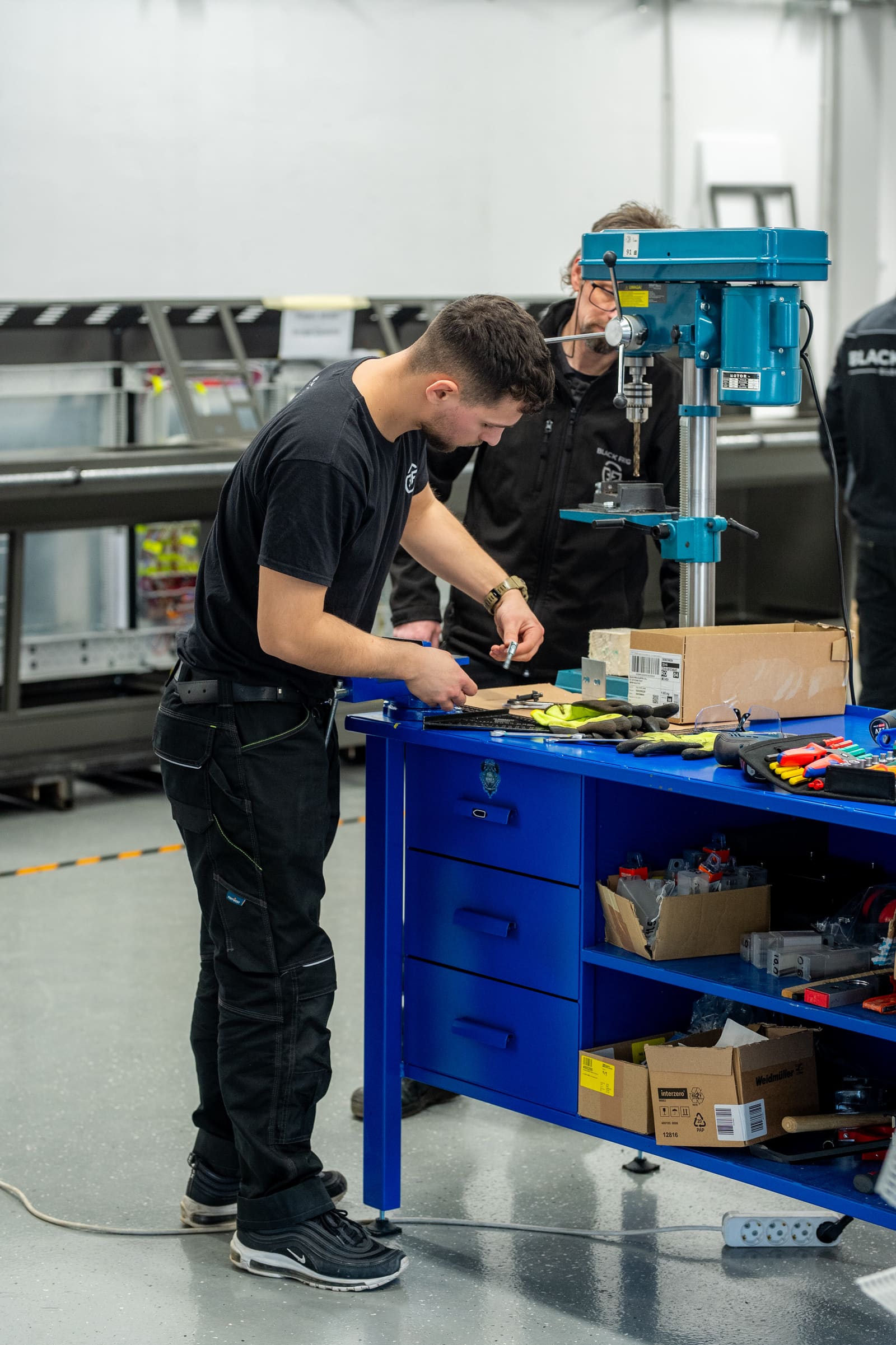 Black Frog Elektro panel builder preparing a component at a workbench while a colleague drills behind him in the workshop