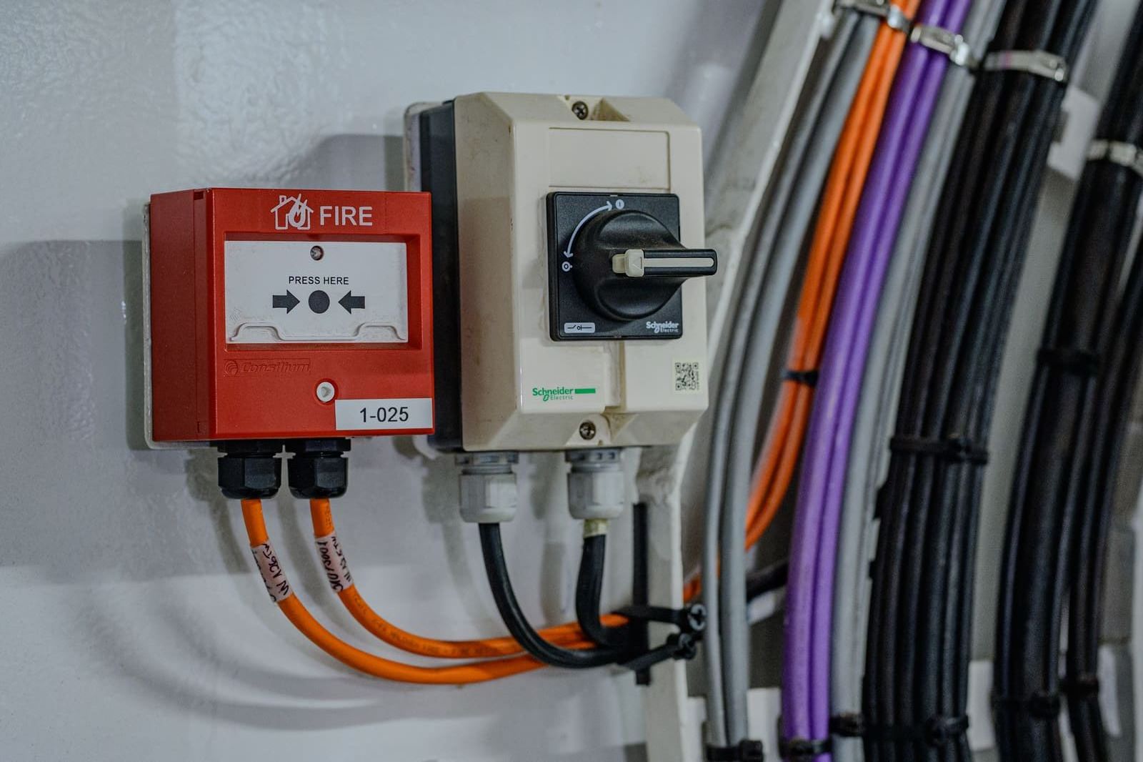 Manual fire-alarm call point and Schneider rotary load switch mounted side by side on a bulkhead next to orange and purple marine cables