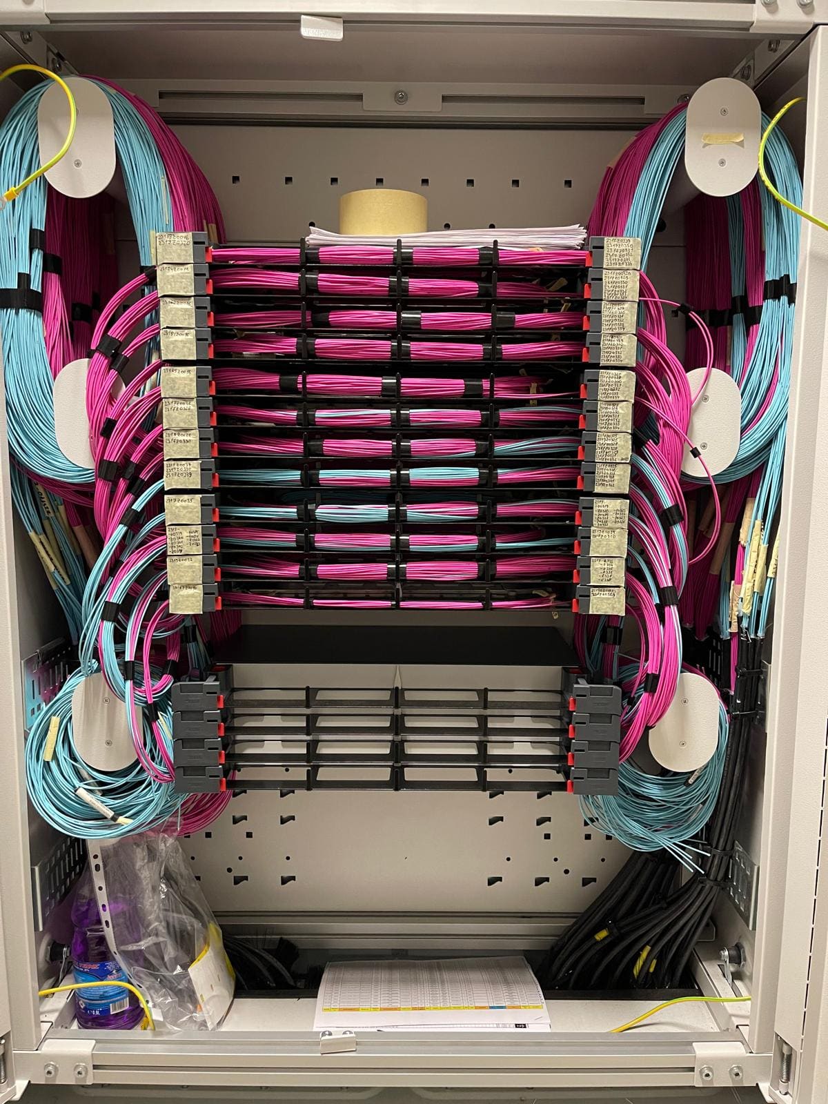 Open fiber optic patch panel with magenta and aqua single-mode and multi-mode patch cords routed to LC adapters across multiple shelves